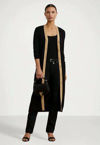 Žena nosi dugi crni kardigan s bež detaljima, crne hlače, crnu majicu, sandale i drži malu crnu torbicu.