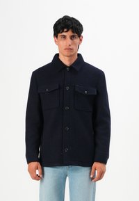 Mann in einer dunkelblauen, geknöpften Jacke mit zwei Brusttaschen, kombiniert mit hellblauen Jeans, steht vor einem schlichten weißen Hintergrund.