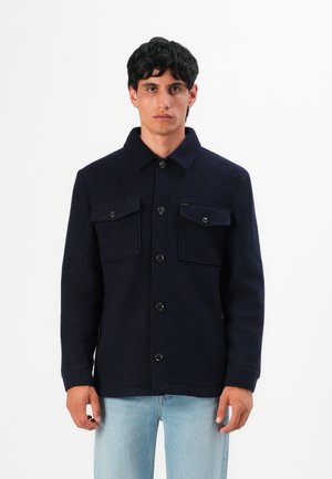 Mann in einer dunkelblauen, geknöpften Jacke mit zwei Brusttaschen, kombiniert mit hellblauen Jeans, steht vor einem schlichten weißen Hintergrund.