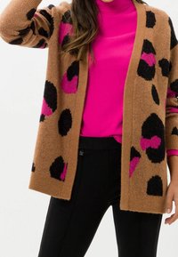 Brun stickad cardigan med rosa och svarta leopardfläckar, öppen framsida, bärs över en ljusrosa polotröja, matchad med svarta byxor.