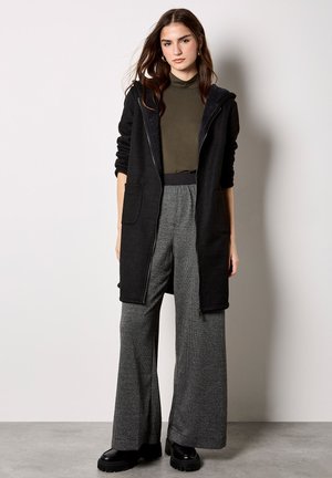 Donna in piedi contro un muro chiaro che indossa un cappotto nero con cappuccio, dolcevita verde oliva, pantaloni grigi a gamba larga e scarpe piattaforma nere.