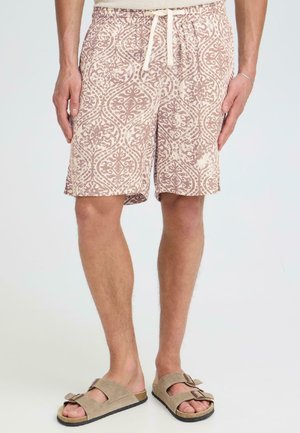 Mand iført beige mønstrede shorts med snøre og beige ruskindssandaler med dobbelte stropper, stående mod en ensfarvet baggrund.