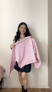 Νεαρή γυναίκα με μακριά σκούρα μαλλιά που κρατάει ένα ανοιχτό ροζ φούτερ, στέκεται μπροστά σε έναν απλό λευκό τοίχο πάνω σε ξύλινο πάτωμα.