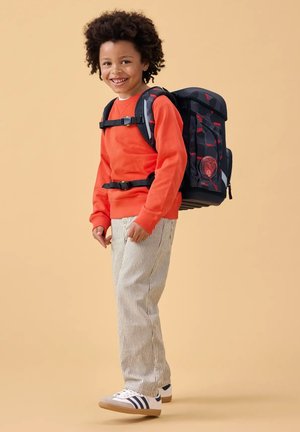 Smiling child with curly hair wearing an orange sweatshirt, striped pants, white sneakers, and a large black and red backpack.