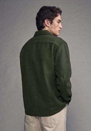 Junger Mann mit dunklem Haar, der ein olivgrünes Langarmhemd und hellbeige Hose trägt, mit dem Rücken zu einem schlichten grauen Hintergrund gewandt.