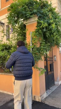 Granatowa kurtka puchowa, beżowe spodnie i kręcone włosy na tle pomarańczowej ściany, zielonej roślinności i czarnej żelaznej bramy.