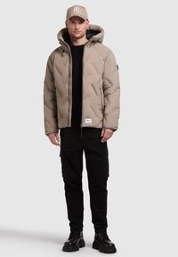 Giacca puffer beige con cappuccio, texture trapuntata, tasche con zip nere, indossata sopra un maglione nero, abbinata a pantaloni cargo neri e stivali.