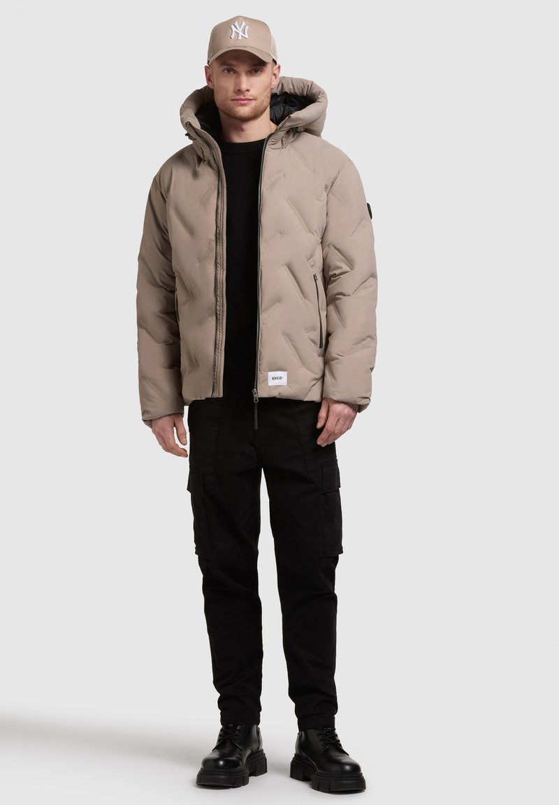 Giacca puffer beige con cappuccio, texture trapuntata, tasche con zip nere, indossata sopra un maglione nero, abbinata a pantaloni cargo neri e stivali.