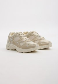 Zapatillas beige con paneles de malla y sintéticos, suela acolchada, diseño de cordones, sutiles acentos blancos y punta redondeada.