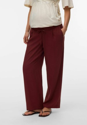 Vrouw draagt comfortabele bordeauxrode broek met trekkoord, beige korte mouwen top met zijkant strik, en zwarte platte sandalen, staand met handen in de zakken.