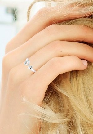 Bague en argent avec un centre en forme de cœur orné de pierres claires, monté sur une bande lisse, portée sur une main avec de longs cheveux clairs.