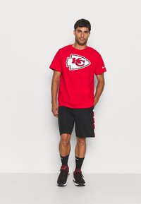 T-shirt rouge à manches courtes avec un logo des Kansas City Chiefs en contour blanc. Porté avec un short noir et des chaussures de sport noir-rouge.