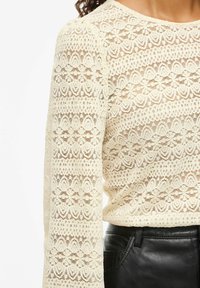 Crème kanten lange mouwen top met ingewikkelde bloemenpatronen, gecombineerd met zwarte leren broek, weergegeven van halverwege de torso tot bovenarm.