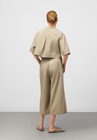 Beige Kurzoberteil mit kurzen Ärmeln und einem Rückenfalten, kombiniert mit lockeren, weit geschnittenen Hosen in einem passenden Farbton, getragen mit einfachen Sandalen.