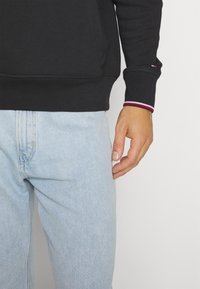 Czarna bluza z ściągaczami w kolorze czerwonym, białym i niebieskim, w zestawieniu z jasnoniebieskimi dżinsami. Ręka opiera się na nodze.