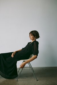 Crna haljina s puf rukavima i teksturiranim materijalom, s lepršavim siluetom. Sjedila na metalnoj stolici pred jednostavnom pozadinom.