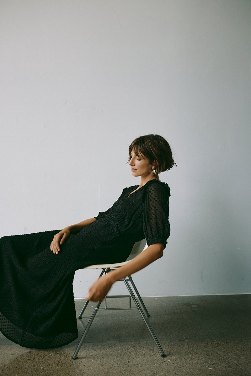 Crna haljina s puf rukavima i teksturiranim materijalom, s lepršavim siluetom. Sjedila na metalnoj stolici pred jednostavnom pozadinom.