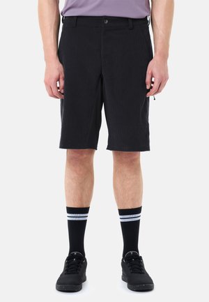 Homme portant un short noir jusqu'aux genoux, des baskets noires et des chaussettes noires avec deux rayures blanches, debout devant un fond clair uni.