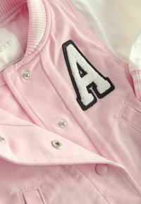 Veste rose avec boutons-pression, écusson lettre A blanc bordé de noir, et col et poignets côtelés visibles à rayures blanches et roses.