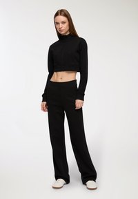 Czarny, krótki sweter z uchwytem na suwak i stójką, z dwiema przednimi kieszeniami, w połączeniu z luźnymi czarnymi spodniami i białymi trampkami.