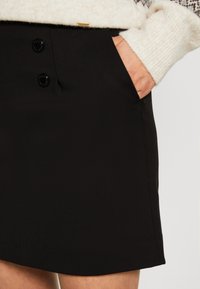 Main dans la poche d'une jupe noire à deux boutons, portée avec un pull en maille crème avec un yoke à motif.