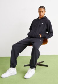 Jauns vīrietis ar pītiem matiem sēž uz brūnas krēsla, ģērbies melnā kapuci, melnos sporta biksēs un baltās Nike kedās uz zaļa grīdas.