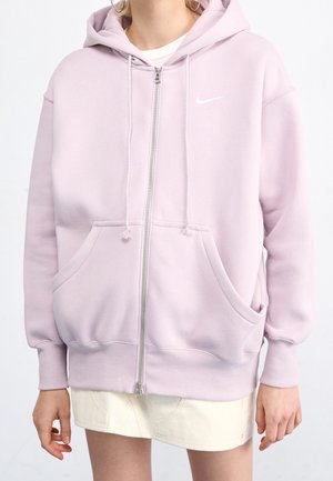Osoba ubrana w jasnofioletową bluzę Nike na zamek z kapturem, sznurkami i przednimi kieszeniami, na białej spódnicy.