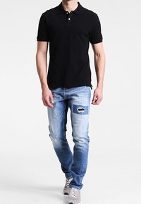 Polo shirt noir à manches courtes, associé à un jean délavé bleu clair avec une pièce bleu foncé et des détails usés. Baskets gris.