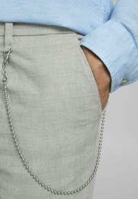 Šedé kostkované kalhoty s kovovým řetězem připevněným k poutku na opasek a viditelným rukávem světle modré texturované košile.