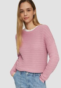 Pull en tricot rose avec un motif texturé, encolure ronde et manches longues, porté par-dessus une chemise blanche, associé à un short en jean.