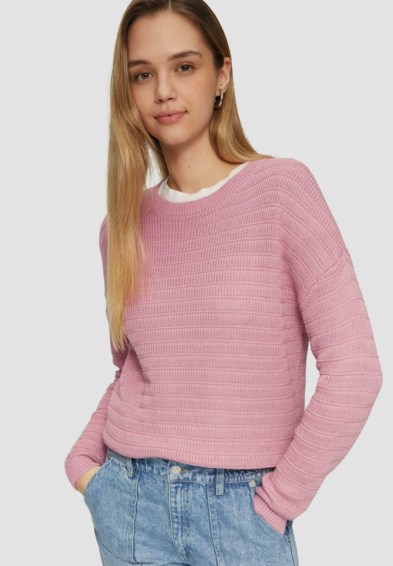 Pull en tricot rose avec un motif texturé, encolure ronde et manches longues, porté par-dessus une chemise blanche, associé à un short en jean.
