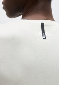 Κοντινή όψη του λαιμού και του πάνω μέρους της πλάτης ενός σκουρόχρωμου ατόμου που φοράει λευκό πουκάμισο με μαύρη κάθετη ετικέτα λογότυπου North Face.