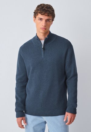 Jonge man met krullend haar die een donkerblauwe trui met een kwart ritssluiting en lichtblauwe spijkerbroek draagt tegen een effen lichte achtergrond.