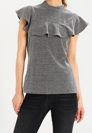Femme portant un haut sans manches gris texturé avec un col et des épaules volantés, associé à un jean noir ajusté sur un fond uni.