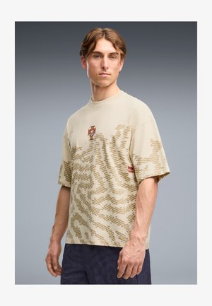 Jeune homme portant une chemise beige à manches courtes à motif avec un blason et un logo, debout devant un fond gris uni.