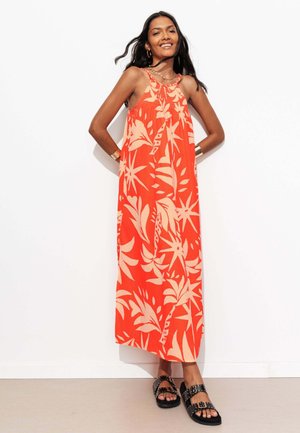 Femme debout souriante portant une robe longue sans manches orange avec des motifs de feuilles tropicales crème et des sandales noires à clous.