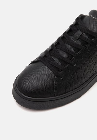 Gros plan d'une basket noire texturée avec des lacets noirs et un motif discret en relief sur le côté, montrant l'avant et une partie de la languette.