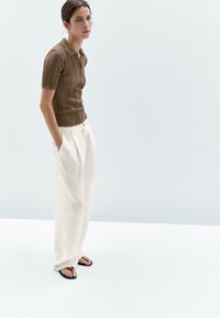 Polo shirt marron côtelé, pantalon blanc à plis larges et tongs noires. Le modèle se tient avec les mains dans les poches contre un fond clair.