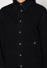 Svart denimskjorta med krage, framknappning i en matchande mörk ton och bröstficka. Har en slät textur och en strukturerad design.