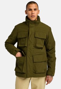 Chaqueta verde oliva con un alto cuello, múltiples bolsillos frontales grandes y una textura suave, diseñada para la utilidad y la calidez.