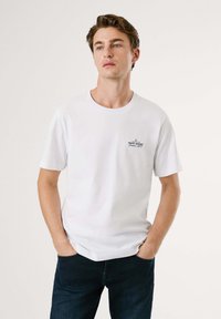 Hvit T-skjorte med korte ermer i bomull og rund hals, med en liten svart logo trykket på venstre brystområde. Brukt med mørke jeans.