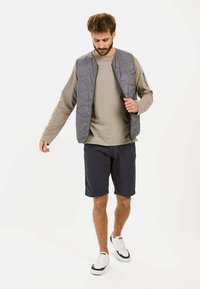 Gilet matelassé gris sur une chemise à manches longues beige, associé à un short bleu marine et des baskets blanches, porté par un mannequin sur fond blanc.