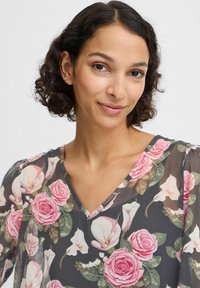 Blomsterbluse med en mørkegrå baggrund, lyserøde roser og hvide kaldeliljer. V-halsdesign med blødt, gennemsigtigt stof og puffede ærmer.