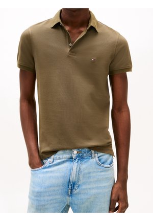 Personne portant un polo à manches courtes vert olive et un jean en denim bleu clair, une main dans la poche, sur un fond uni.