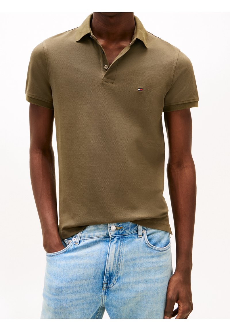 Personne portant un polo à manches courtes vert olive et un jean en denim bleu clair, une main dans la poche, sur un fond uni.
