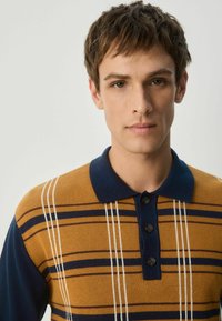 Polo-shirt i sennepsfarve med marineblå accenter, prydet med horisontale striber og tre sorte knapper ved kraven. Blød strikket tekstur.