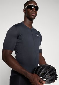 Maillot de cyclisme bleu marine foncé avec une finition texturée, à manches courtes et fermeture éclair sur le devant. Des mains tenant un casque de vélo noir.