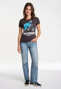 Schwarzes T-Shirt mit blauem grafischen Design eines Charakters auf einem Motorrad, kombiniert mit hellblauen Jeans und dunklen Sneakern. Einfacher, lässiger Schnitt.