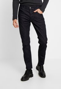 Jeans de mezclilla oscura con un corte ajustado, que cuentan con cinco bolsillos, cierre de botón y una ligera textura en la superficie. Combinados con zapatos negros de cordones.