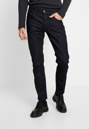 Jeans de mezclilla oscura con un corte ajustado, que cuentan con cinco bolsillos, cierre de botón y una ligera textura en la superficie. Combinados con zapatos negros de cordones.